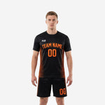 Custom men's soccer team uniforms in jet black with athletic orange stripe panels, front view showing arched team name and number on jersey and matching shorts