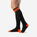 Custom orange and black soccer socks three-quarter angle showing orange toe panel and full calf length fit on adult male model