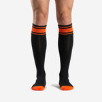 Custom orange and black soccer socks front view showing double orange stripe cuff band and jet black calf body on adult male model