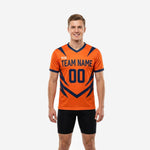 Custom orange soccer training jersey with navy blue geometric strike graphics, team name and number on chest, V-neckline, front view on male model